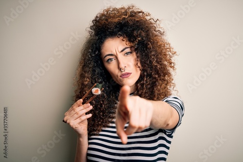 Young beautiful woman with curly hair and piercing eating sushi using wooden chopsticks pointing with finger to the camera and to you, hand sign, positive and confident gesture from the front
