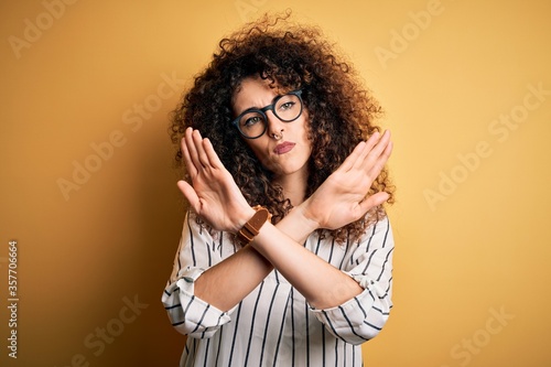 Young beautiful woman with curly hair and piercing wearing striped shirt and glasses Rejection expression crossing arms doing negative sign, angry face