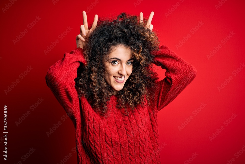 Young beautiful woman with curly hair and piercing wearing casual red sweater Posing funny and crazy with fingers on head as bunny ears, smiling cheerful Young beautiful woman with curly hair and piercing wearing casual red sweater Posing funny and crazy with fingers on head as bunny ears, smiling cheerful