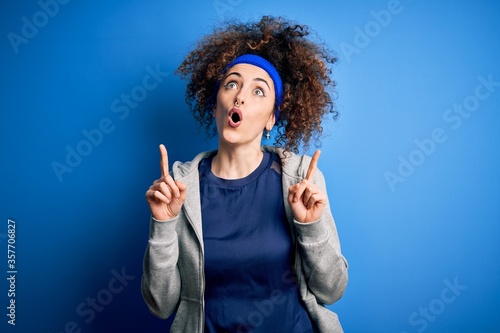 Young beautiful sportswoman with curly hair and piercing standing wearing sportswear amazed and surprised looking up and pointing with fingers and raised arms.