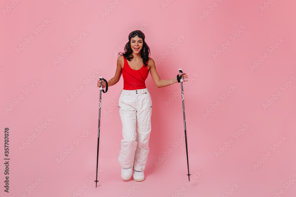 Smiling adorable woman posing with ski poles. Full length view of girl in white pants standing on pink background. Smiling adorable woman posing with ski poles. Full length view of girl in white pants standing on pink background.