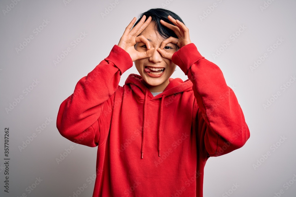 Young beautiful asian girl wearing casual sweatshirt with hoodie over white background doing ok gesture like binoculars sticking tongue out, eyes looking through fingers. Crazy expression. Young beautiful asian girl wearing casual sweatshirt with hoodie over white background doing ok gesture like binoculars sticking tongue out, eyes looking through fingers. Crazy expression.