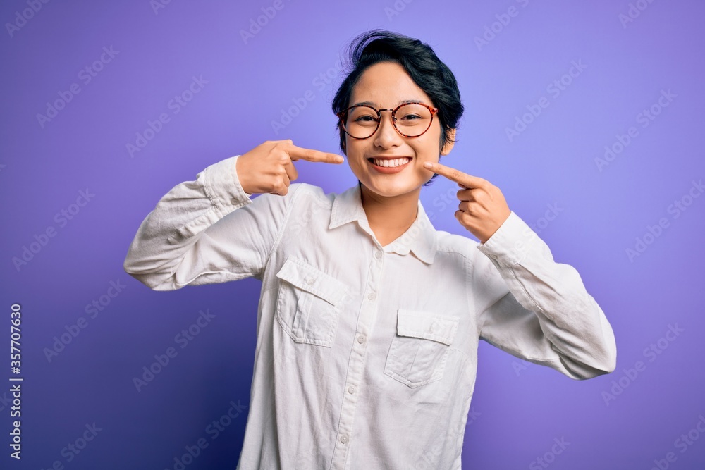 Young beautiful asian girl wearing casual shirt and glasses standing over purple background smiling cheerful showing and pointing with fingers teeth and mouth. Dental health concept. Young beautiful asian girl wearing casual shirt and glasses standing over purple background smiling cheerful showing and pointing with fingers teeth and mouth. Dental health concept.