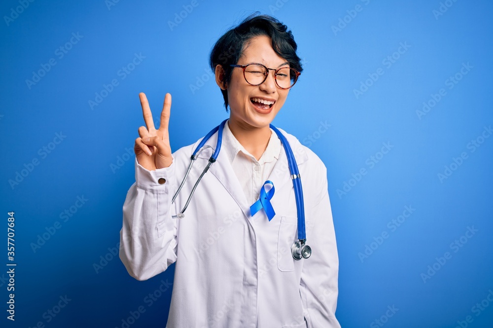 Young beautiful asian doctor girl wearing stethoscope and coat with blue cancer ribbon smiling with happy face winking at the camera doing victory sign. Number two. Young beautiful asian doctor girl wearing stethoscope and coat with blue cancer ribbon smiling with happy face winking at the camera doing victory sign. Number two.