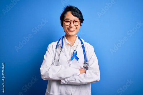 Young beautiful asian doctor girl wearing stethoscope and coat with blue cancer ribbon happy face smiling with crossed arms looking at the camera. Positive person.