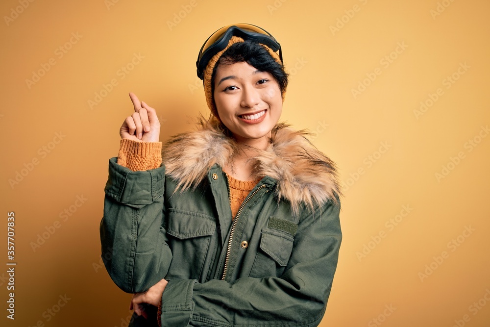Beautiful asian skier girl wearing snow sportswear using ski goggles over yellow background with a big smile on face, pointing with hand and finger to the side looking at the camera. Beautiful asian skier girl wearing snow sportswear using ski goggles over yellow background with a big smile on face, pointing with hand and finger to the side looking at the camera.