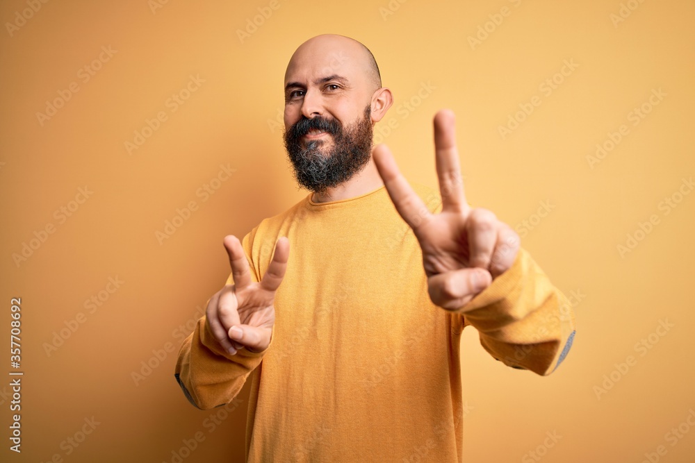 Handsome bald man with beard wearing casual sweater standing over yellow background smiling looking to the camera showing fingers doing victory sign. Number two. Handsome bald man with beard wearing casual sweater standing over yellow background smiling looking to the camera showing fingers doing victory sign. Number two.