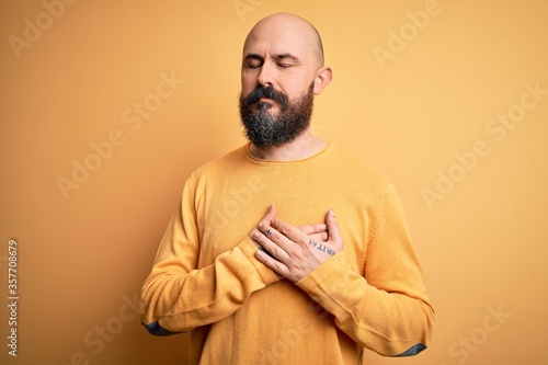 Handsome bald man with beard wearing casual sweater standing over yellow background smiling with hands on chest with closed eyes and grateful gesture on face. Health concept. © Krakenimages.com
