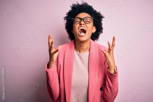 Young beautiful African American afro businesswoman with curly hair wearing pink jacket celebrating mad and crazy for success with arms raised and closed eyes screaming excited. Winner concept