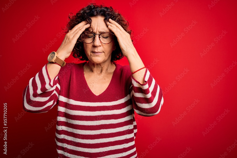 Middle age beautiful curly hair woman wearing casual striped sweater over red background with hand on head for pain in head because stress. Suffering migraine. Middle age beautiful curly hair woman wearing casual striped sweater over red background with hand on head for pain in head because stress. Suffering migraine.