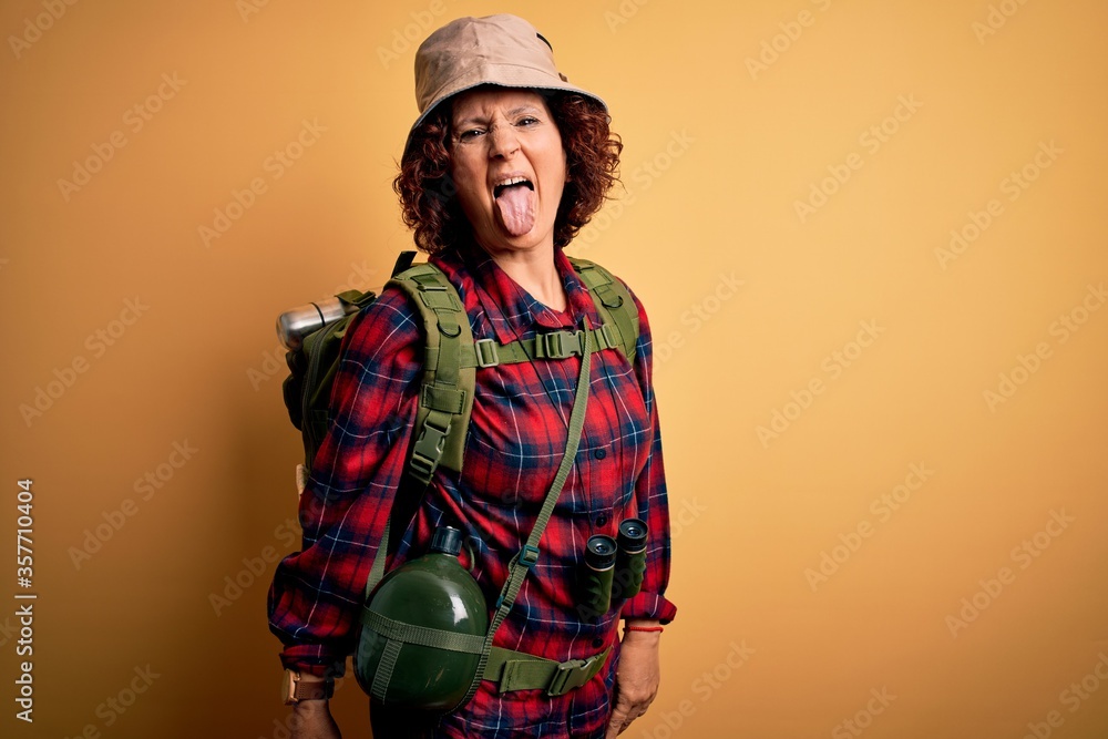Middle age curly hair hiker woman hiking wearing backpack and water canteen using binoculars sticking tongue out happy with funny expression. Emotion concept.