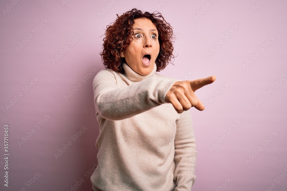 Middle age beautiful curly hair woman wearing casual turtleneck sweater over pink background Pointing with finger surprised ahead, open mouth amazed expression, something on the front Middle age beautiful curly hair woman wearing casual turtleneck sweater over pink background Pointing with finger surprised ahead, open mouth amazed expression, something on the front