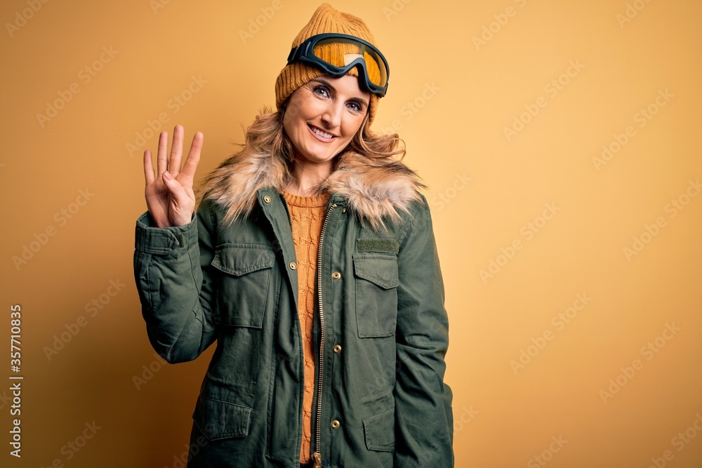 Middle age beautiful blonde skier woman wearing snow sportwear and ski goggles showing and pointing up with fingers number four while smiling confident and happy.