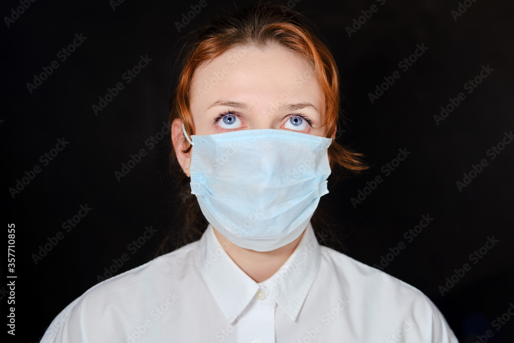Woman in a medical mask who is looking up in thought. Redhead girl raised her eyes up on a black background. Concept of challenges with the coronavirus epidemic Woman in a medical mask who is looking up in thought. Redhead girl raised her eyes up on a black background. Concept of challenges with the coronavirus epidemic
