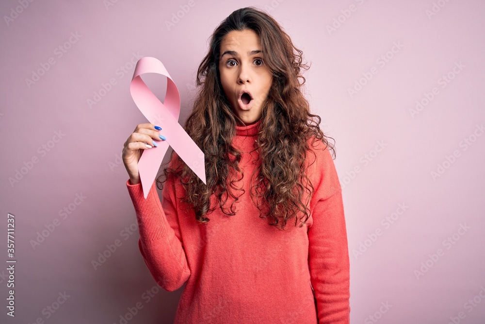 Young beautiful woman with curly hair holding pink cancer ribbon over isolated background scared in shock with a surprise face, afraid and excited with fear expression Young beautiful woman with curly hair holding pink cancer ribbon over isolated background scared in shock with a surprise face, afraid and excited with fear expression