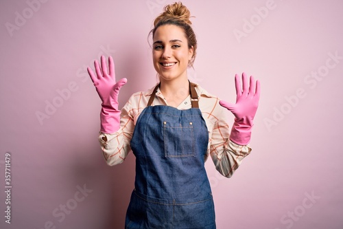 Young beautiful blonde cleaner woman doing housework wearing arpon and gloves showing and pointing up with fingers number ten while smiling confident and happy.