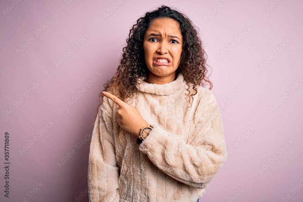 Young beautiful woman with curly hair wearing casual sweater standing over pink background Pointing aside worried and nervous with forefinger, concerned and surprised expression Young beautiful woman with curly hair wearing casual sweater standing over pink background Pointing aside worried and nervous with forefinger, concerned and surprised expression