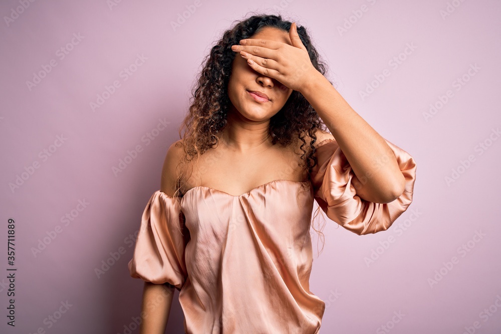 Fototapeta premium Young beautiful woman with curly hair wearing casual t-shirt standing over pink background covering eyes with hand, looking serious and sad. Sightless, hiding and rejection concept