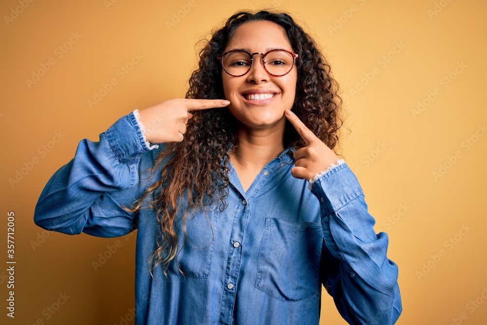 Beautiful woman with curly hair wearing casual denim shirt and glasses over yellow background smiling cheerful showing and pointing with fingers teeth and mouth. Dental health concept. Beautiful woman with curly hair wearing casual denim shirt and glasses over yellow background smiling cheerful showing and pointing with fingers teeth and mouth. Dental health concept.
