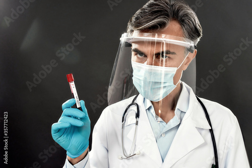 Serious mature male doctor in face shield and blue medical gloves holding positive blood test result for COVID-19, Coronavirus. Looking at camera