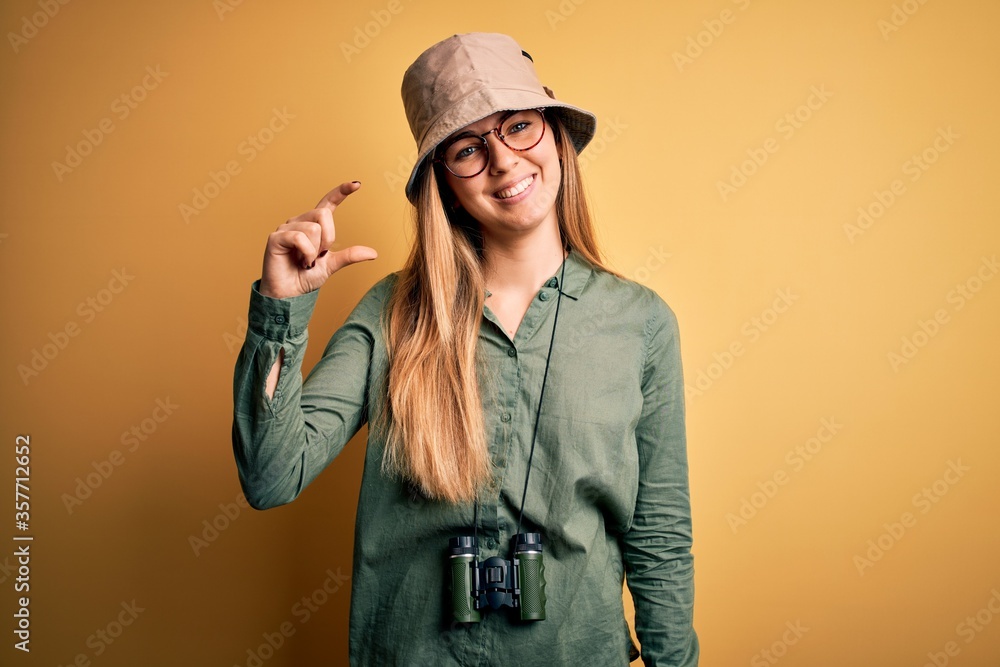 Beautiful blonde explorer woman with blue eyes wearing hat and glasses using binoculars smiling and confident gesturing with hand doing small size sign with fingers looking and the camera. Measure. Beautiful blonde explorer woman with blue eyes wearing hat and glasses using binoculars smiling and confident gesturing with hand doing small size sign with fingers looking and the camera. Measure.