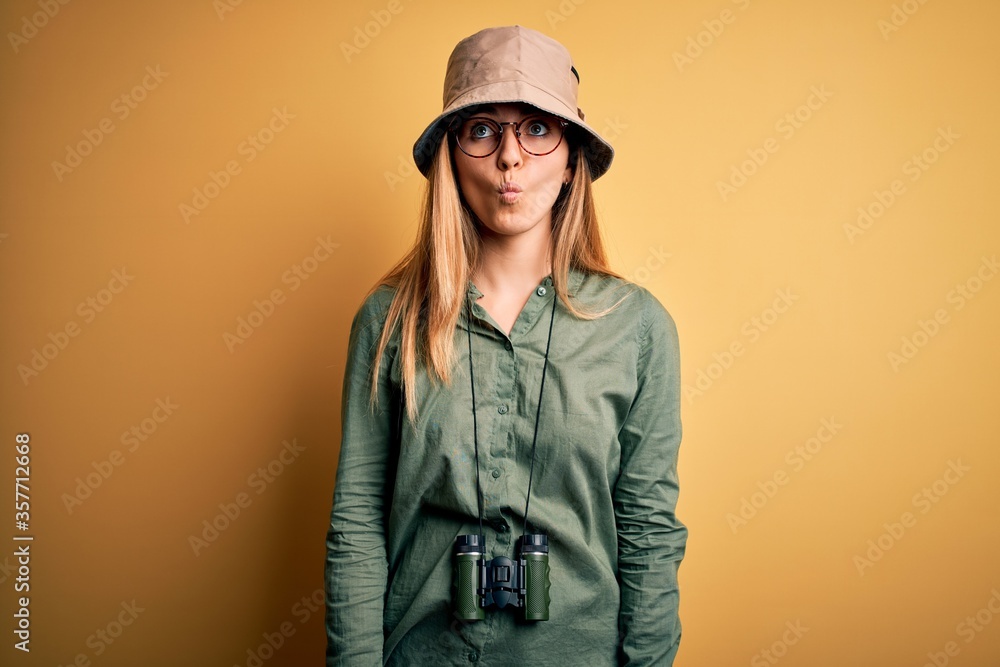 Beautiful blonde explorer woman with blue eyes wearing hat and glasses using binoculars making fish face with lips, crazy and comical gesture. Funny expression.