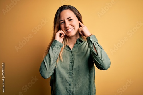 Young beautiful blonde woman with blue eyes wearing green shirt over yellow background covering ears with fingers with annoyed expression for the noise of loud music. Deaf concept.