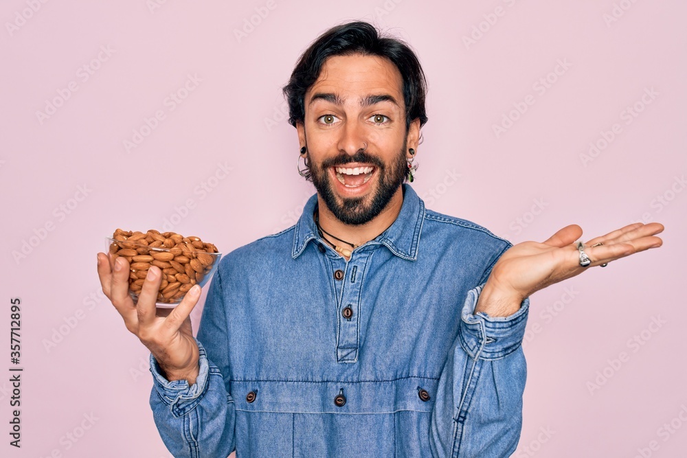 Young handsome hispanic man with bear holding natural ingredient healthy almonds very happy and excited, winner expression celebrating victory screaming with big smile and raised hands Young handsome hispanic man with bear holding natural ingredient healthy almonds very happy and excited, winner expression celebrating victory screaming with big smile and raised hands