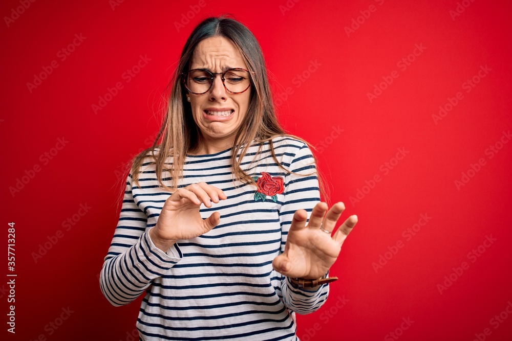 Young beautiful blonde woman with blue eyes wearing glasses standing over red background disgusted expression, displeased and fearful doing disgust face because aversion reaction. Young beautiful blonde woman with blue eyes wearing glasses standing over red background disgusted expression, displeased and fearful doing disgust face because aversion reaction.