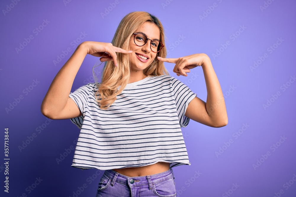 Young beautiful blonde woman wearing striped t-shirt and glasses over purple background smiling cheerful showing and pointing with fingers teeth and mouth. Dental health concept. Young beautiful blonde woman wearing striped t-shirt and glasses over purple background smiling cheerful showing and pointing with fingers teeth and mouth. Dental health concept.
