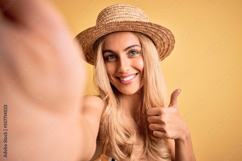 Young beautiful blonde woman on vacation wearing bikini and hat making selfie by camera happy with big smile doing ok sign, thumb up with fingers, excellent sign