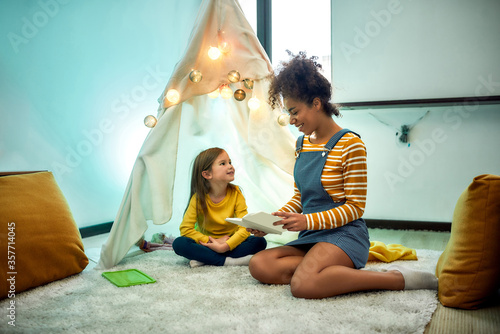Where happiness rules. African american woman baby sitter entertaining little girl. Kid showing a book to her nanny sitting in wigwam, tent. Children education, leisure activity, babysitting concept