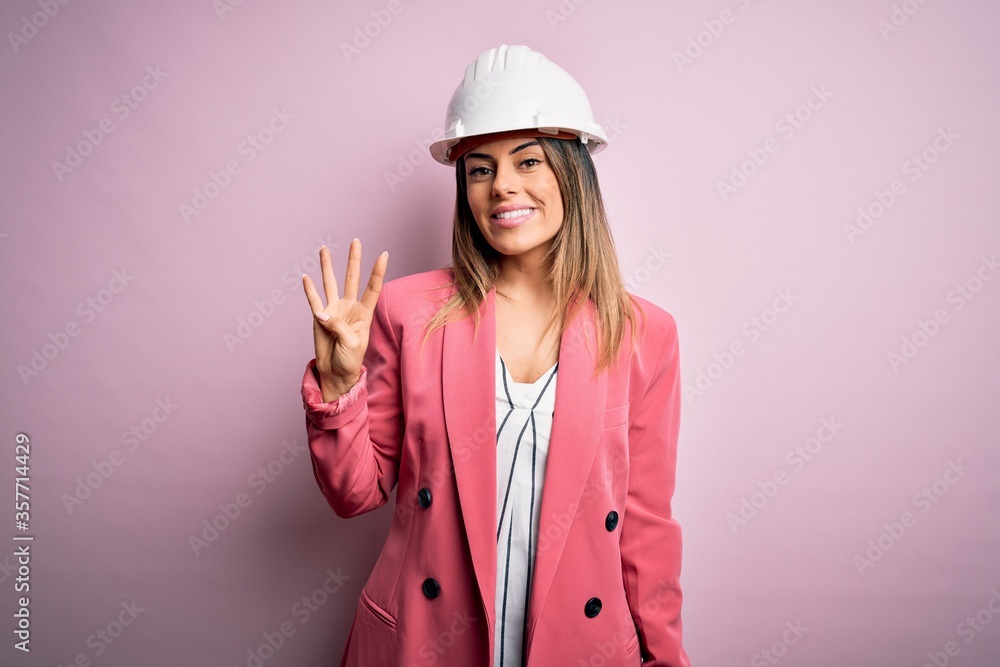 Young beautiful brunette architect woman wearing safety helmet over pink background showing and pointing up with fingers number four while smiling confident and happy.