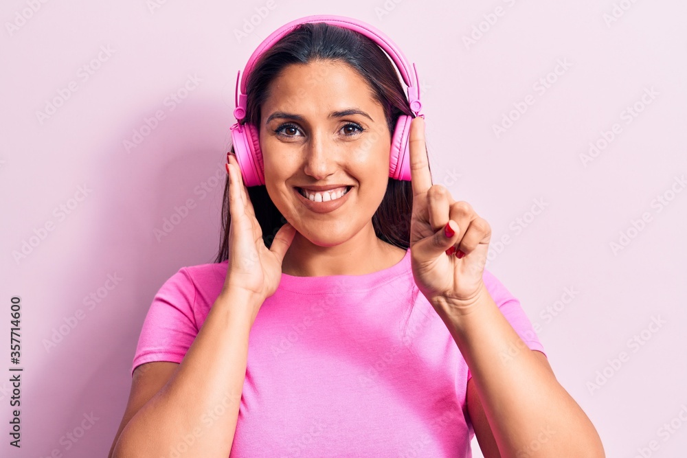 Young beautiful brunette woman listening to music using headphones smiling with an idea or question pointing finger with happy face, number one
