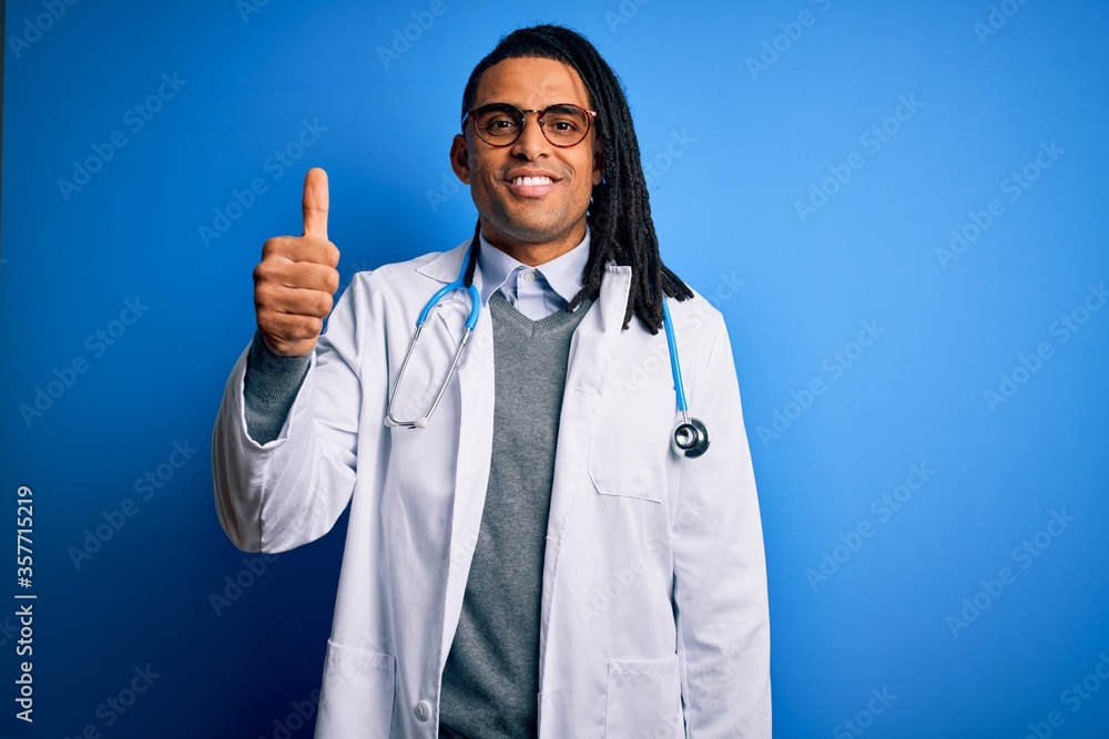 Young african american doctor man with dreadlocks wearing stethoscope and glasses doing happy thumbs up gesture with hand. Approving expression looking at the camera showing success. Young african american doctor man with dreadlocks wearing stethoscope and glasses doing happy thumbs up gesture with hand. Approving expression looking at the camera showing success.