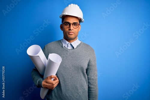 Young african american architect man wearing safety helmet holding bluprints with a confident expression on smart face thinking serious photo