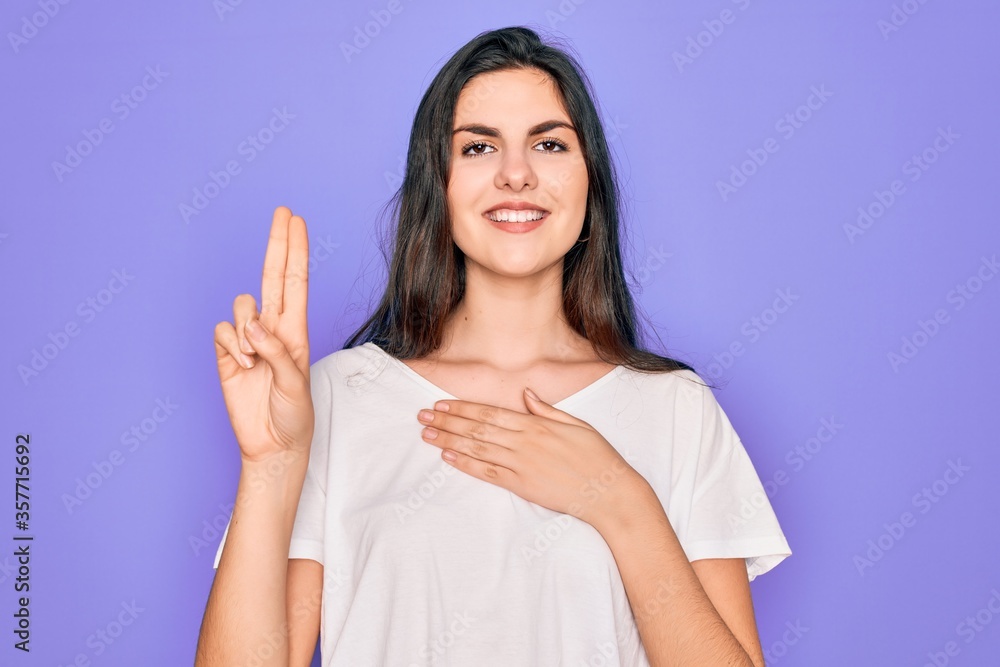 Young beautiful brunette woman wearing casual white t-shirt over purple background smiling swearing with hand on chest and fingers up, making a loyalty promise oath Young beautiful brunette woman wearing casual white t-shirt over purple background smiling swearing with hand on chest and fingers up, making a loyalty promise oath