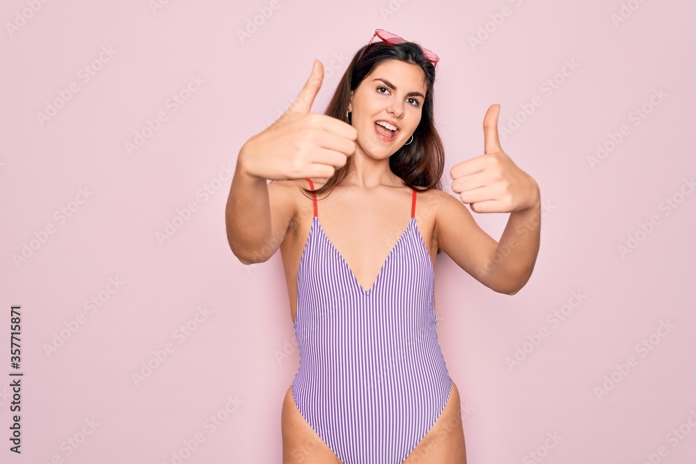 Young beautiful fashion girl wearing swimwear swimsuit and sunglasses over pink background approving doing positive gesture with hand, thumbs up smiling and happy for success. Winner gesture. Young beautiful fashion girl wearing swimwear swimsuit and sunglasses over pink background approving doing positive gesture with hand, thumbs up smiling and happy for success. Winner gesture.