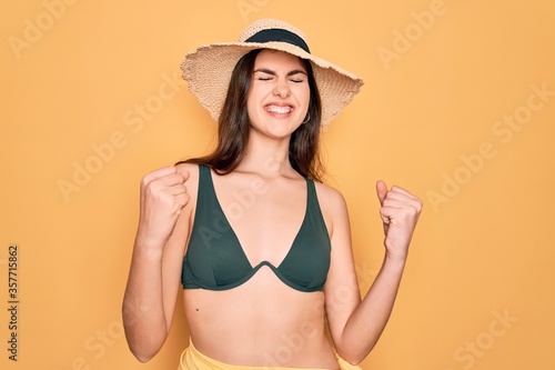 Young beautiful girl wearing swimwear bikini and summer sun hat over yellow background excited for success with arms raised and eyes closed celebrating victory smiling. Winner concept.