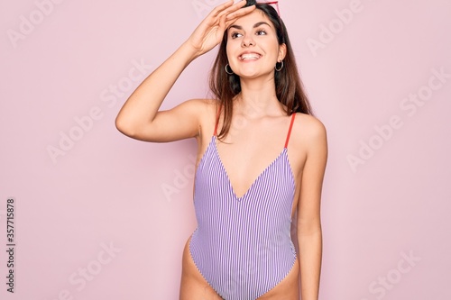 Young beautiful fashion girl wearing swimwear swimsuit and sunglasses over pink background very happy and smiling looking far away with hand over head. Searching concept.