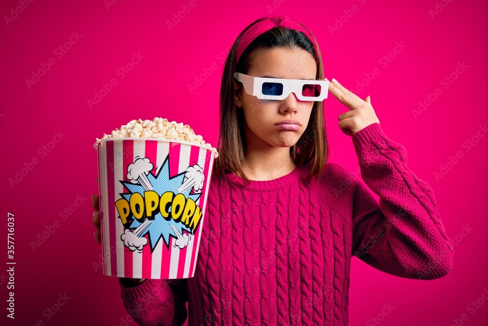 Obraz premium Young beautiful girl watching movie using 3d glasses eating box with popcorns Shooting and killing oneself pointing hand and fingers to head like gun, suicide gesture.