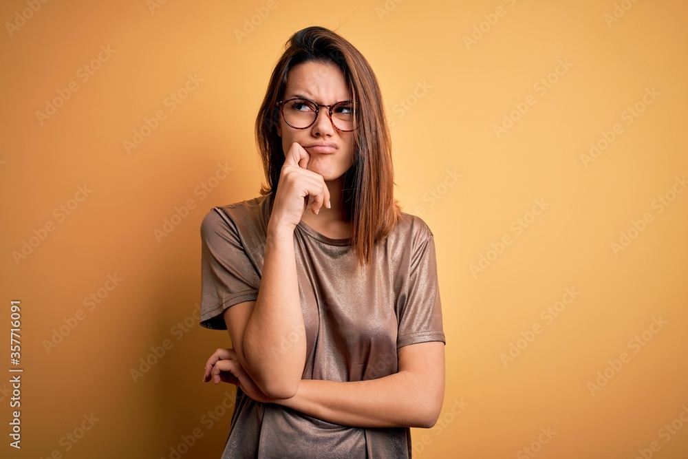 Beautiful brunette girl wearing casual t-shirt and glasses over isolated yellow background with hand on chin thinking about question, pensive expression. Smiling with thoughtful face. Doubt concept. Beautiful brunette girl wearing casual t-shirt and glasses over isolated yellow background with hand on chin thinking about question, pensive expression. Smiling with thoughtful face. Doubt concept.