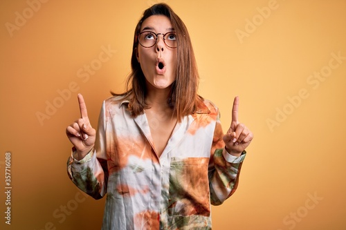 Young beautiful brunette girl wearing casual shirt and glasses over isolated yellow background amazed and surprised looking up and pointing with fingers and raised arms.