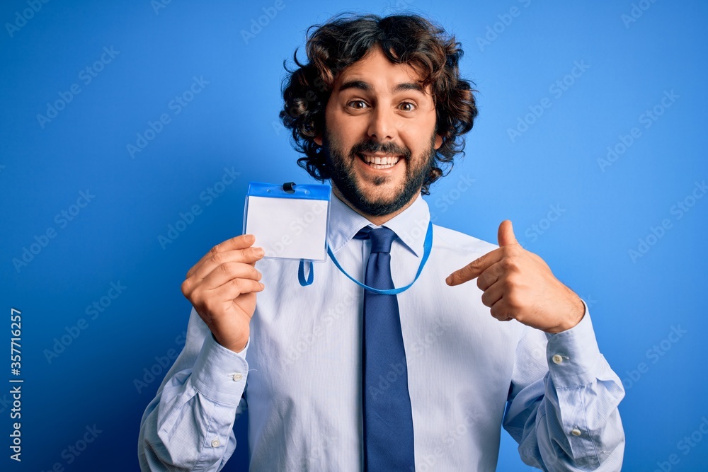 Young handsome business man with beard holding id card identification over blue background with surprise face pointing finger to himself Young handsome business man with beard holding id card identification over blue background with surprise face pointing finger to himself