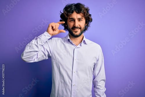 Young handsome business man with beard wearing shirt standing over purple background smiling and confident gesturing with hand doing small size sign with fingers looking and the camera. Measure.