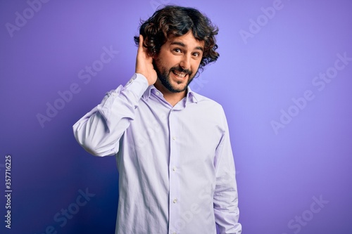 Young handsome business man with beard wearing shirt standing over purple background smiling with hand over ear listening an hearing to rumor or gossip. Deafness concept.