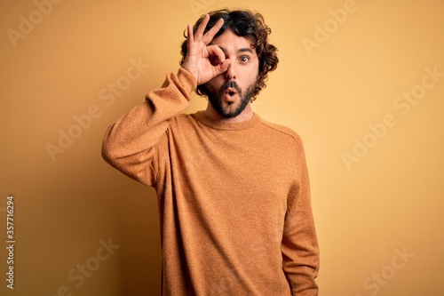 Young handsome man with beard wearing casual sweater standing over yellow background doing ok gesture shocked with surprised face, eye looking through fingers. Unbelieving expression.