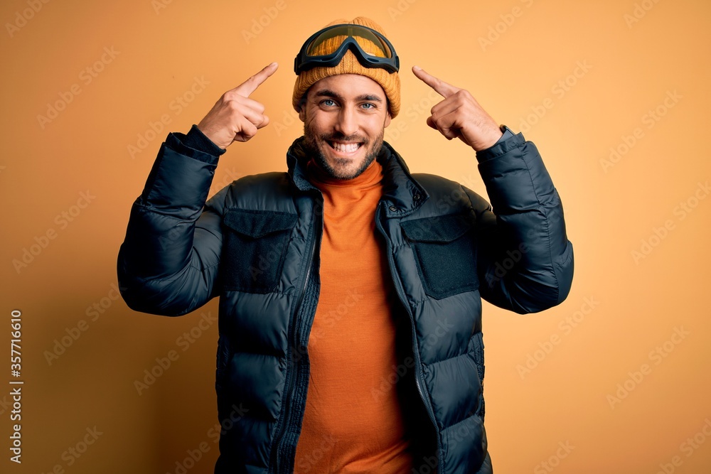 Young handsome skier man with beard wearing snow sportswear and ski goggles smiling pointing to head with both hands finger, great idea or thought, good memory Young handsome skier man with beard wearing snow sportswear and ski goggles smiling pointing to head with both hands finger, great idea or thought, good memory