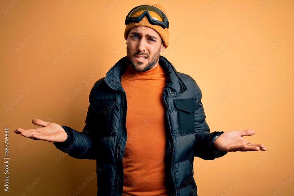 Young handsome skier man with beard wearing snow sportswear and ski goggles clueless and confused with open arms, no idea concept. Young handsome skier man with beard wearing snow sportswear and ski goggles clueless and confused with open arms, no idea concept.