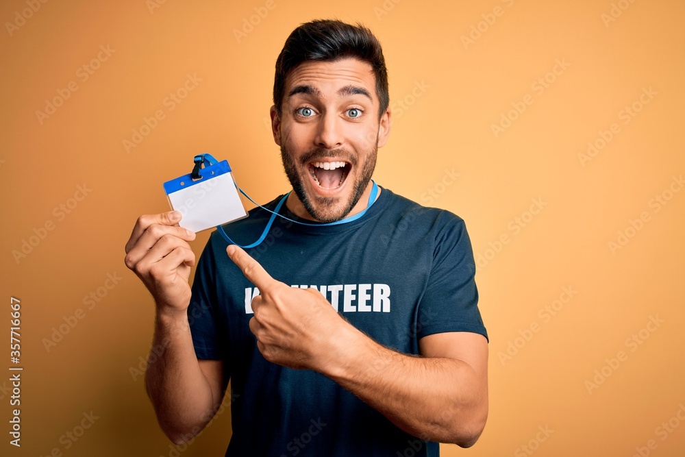 Handsome volunteer man with beard holding id card identification over yellow background very happy pointing with hand and finger Handsome volunteer man with beard holding id card identification over yellow background very happy pointing with hand and finger